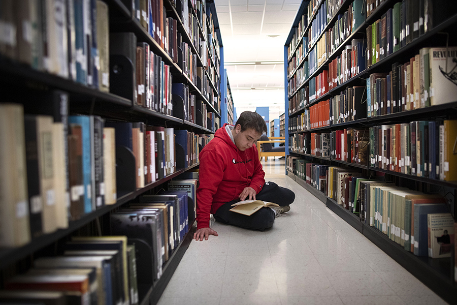 A student sits on the floor of a library and reads.