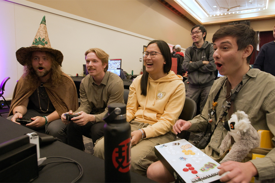 Four people gather around a gaming system and react enthusiastically to what's on the screen.
