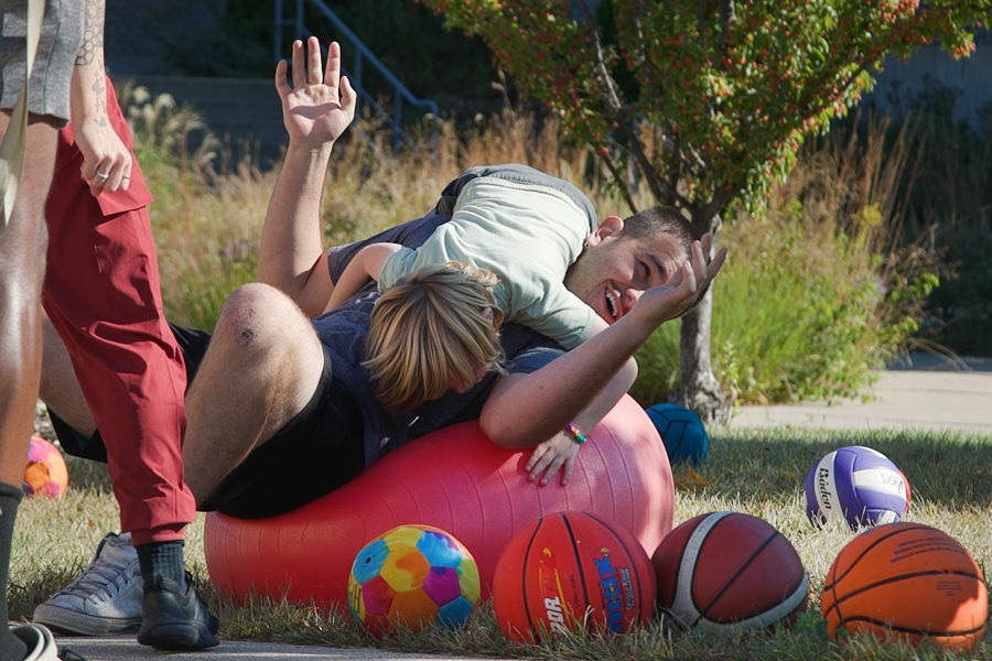 A student lays on a large red exercise ball and laughs as a child jumps on him.