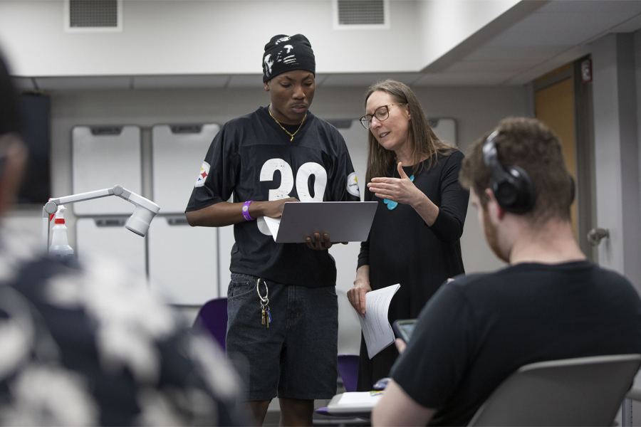 A student stands and holds a laptop while the faculty member looks on.